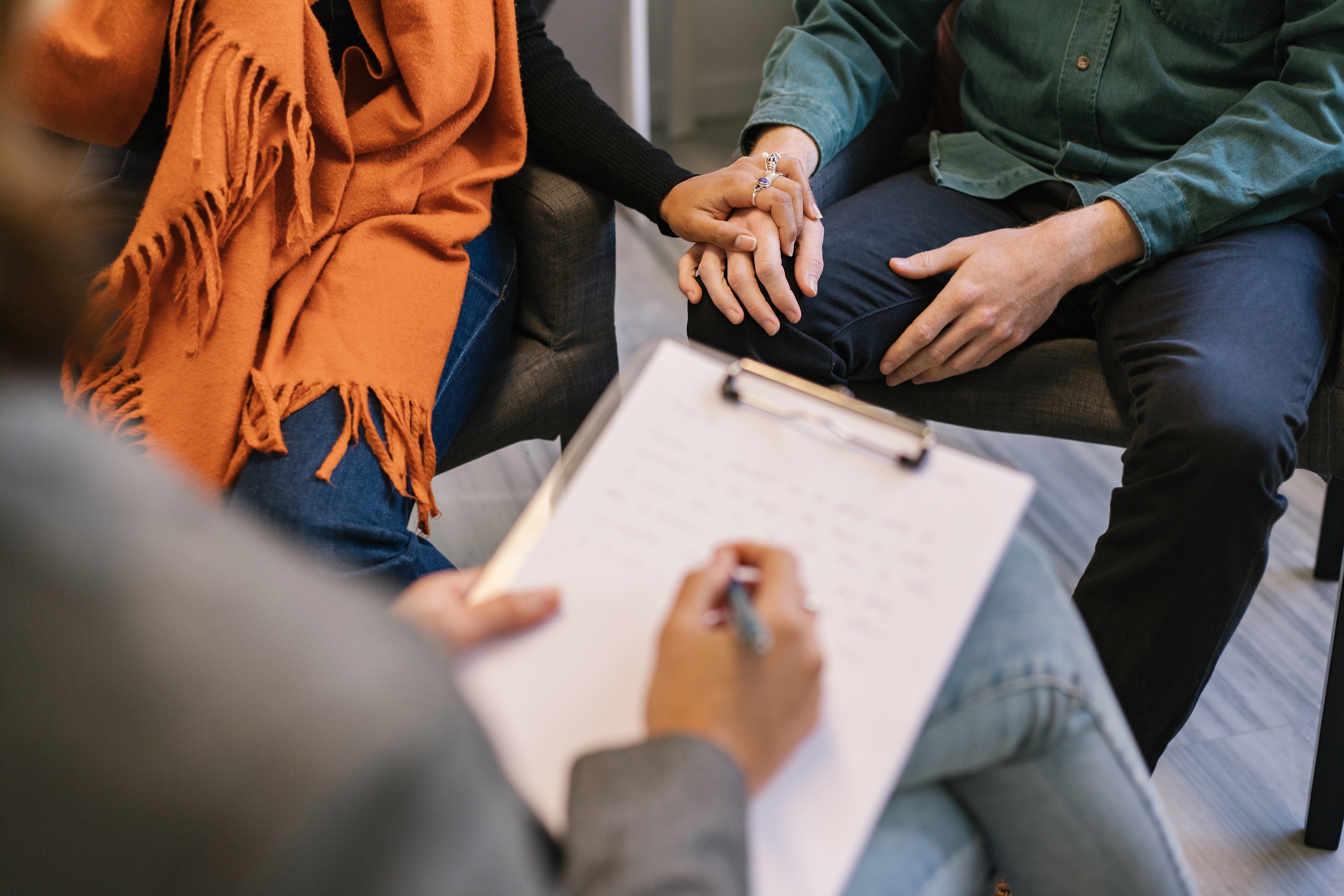 Two people holding hands in a therapy session, with a therapist writing on a clipboard in the foreground