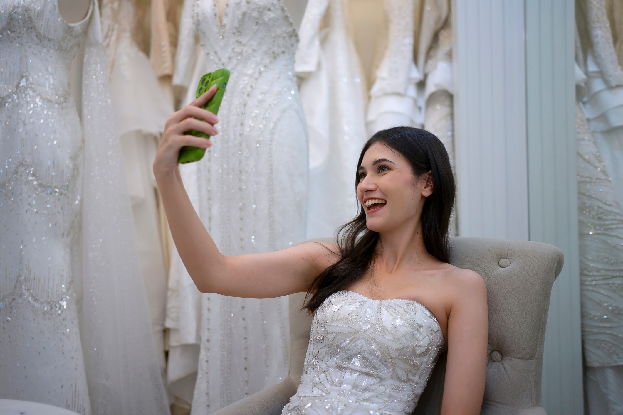 A woman in an elegant strapless gown takes a selfie, seated amidst several hanging bridal dresses in a boutique