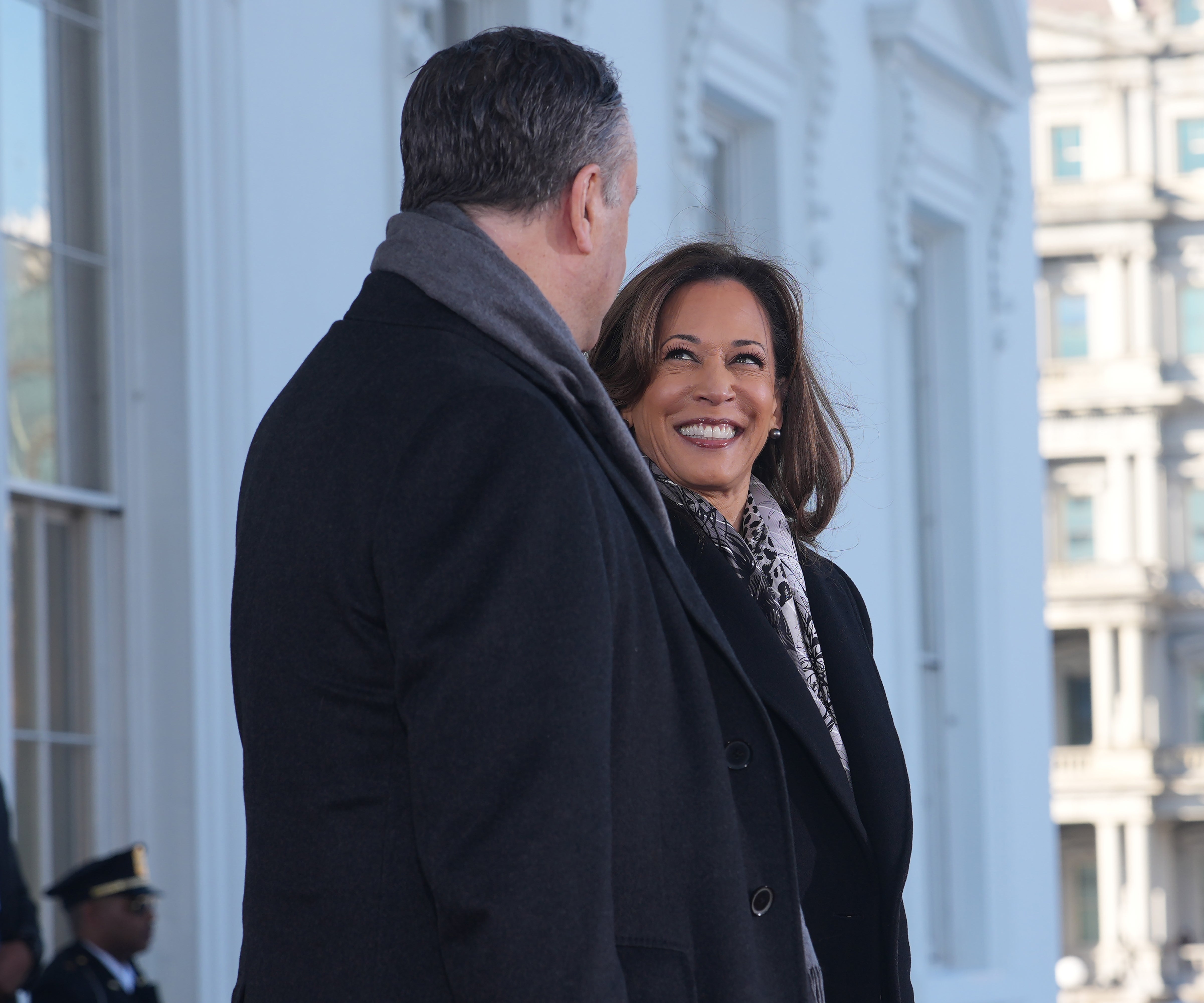 Two people stand outdoors, smiling at each other. One is wearing a dark coat and patterned scarf; the other is in a dark coat with a scarf