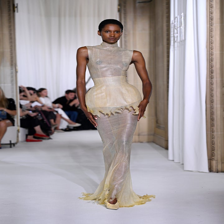 Model walks runway in a form-fitting, sheer gown with a structured, ruffled peplum detail. Audience seated along the side