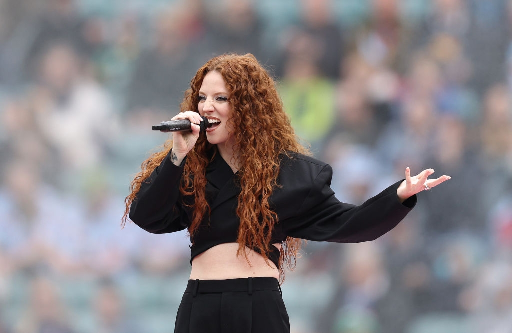 Person sings passionately into a microphone, wearing a stylish black outfit with long sleeves and a cropped top