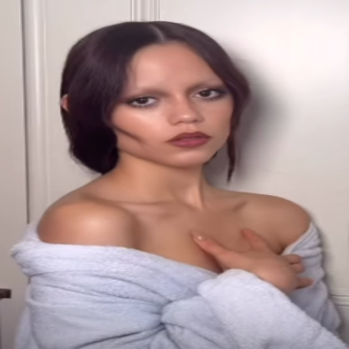 Person with long hair, wearing a robe, stands indoors with a hand on their chest, gazing at the camera