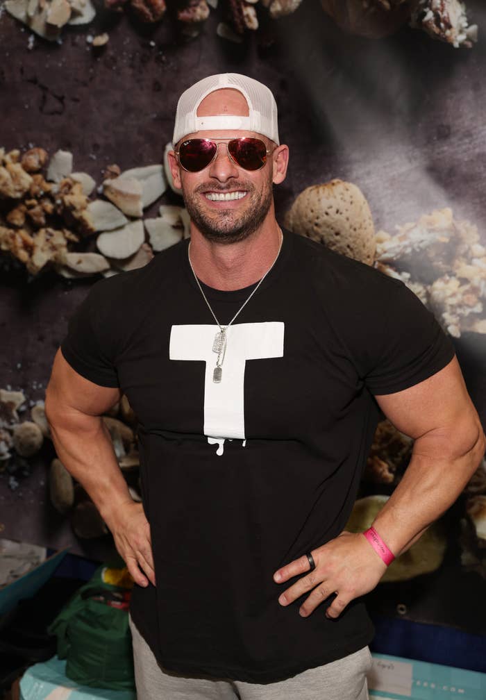 Man wearing a T-shirt, sunglasses, and a baseball cap, posing with hands on hips. Background features a textured design