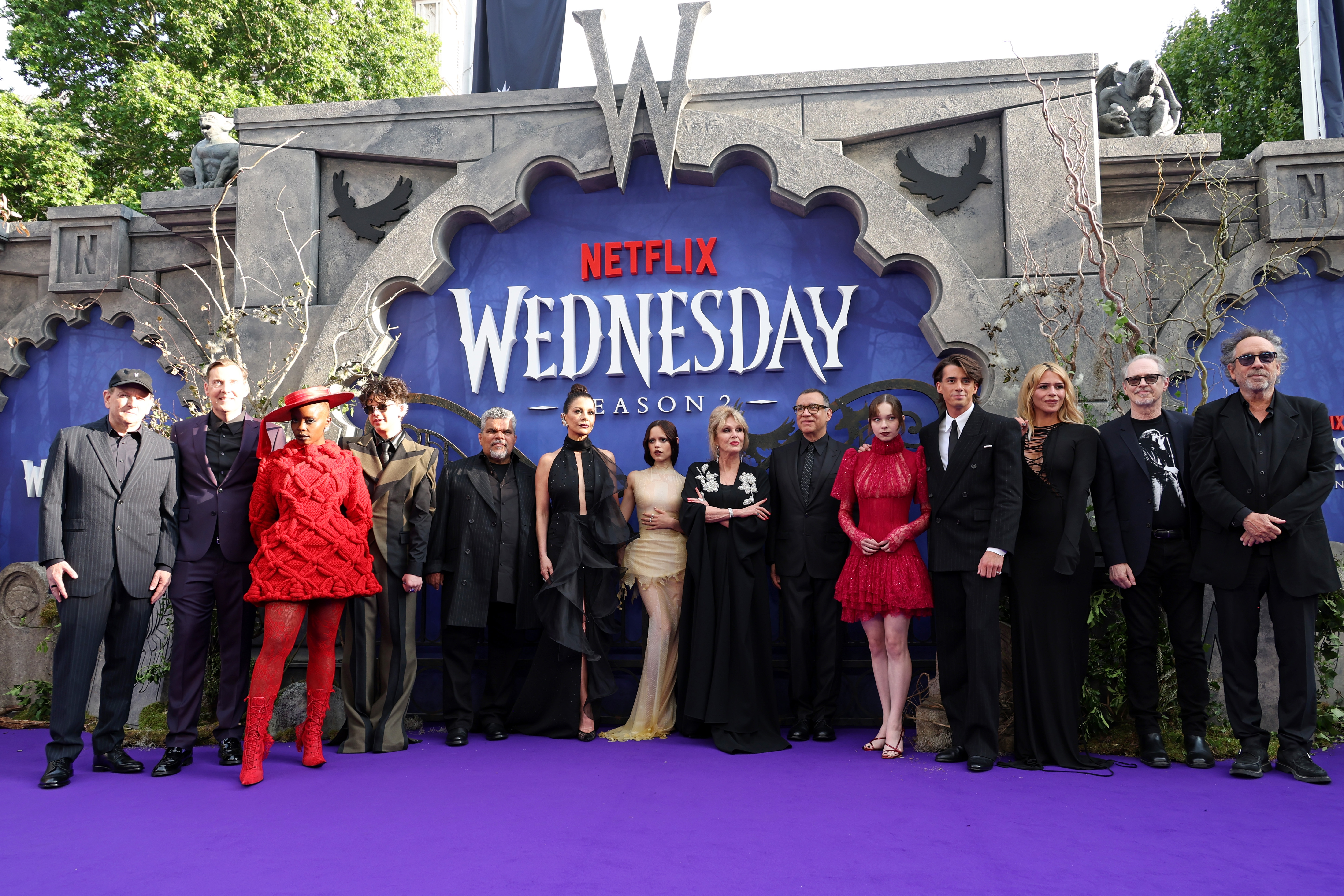 A group of people poses at a "Wednesday" event. They are dressed in formal, eclectic styles, standing on a purple carpet with a themed backdrop