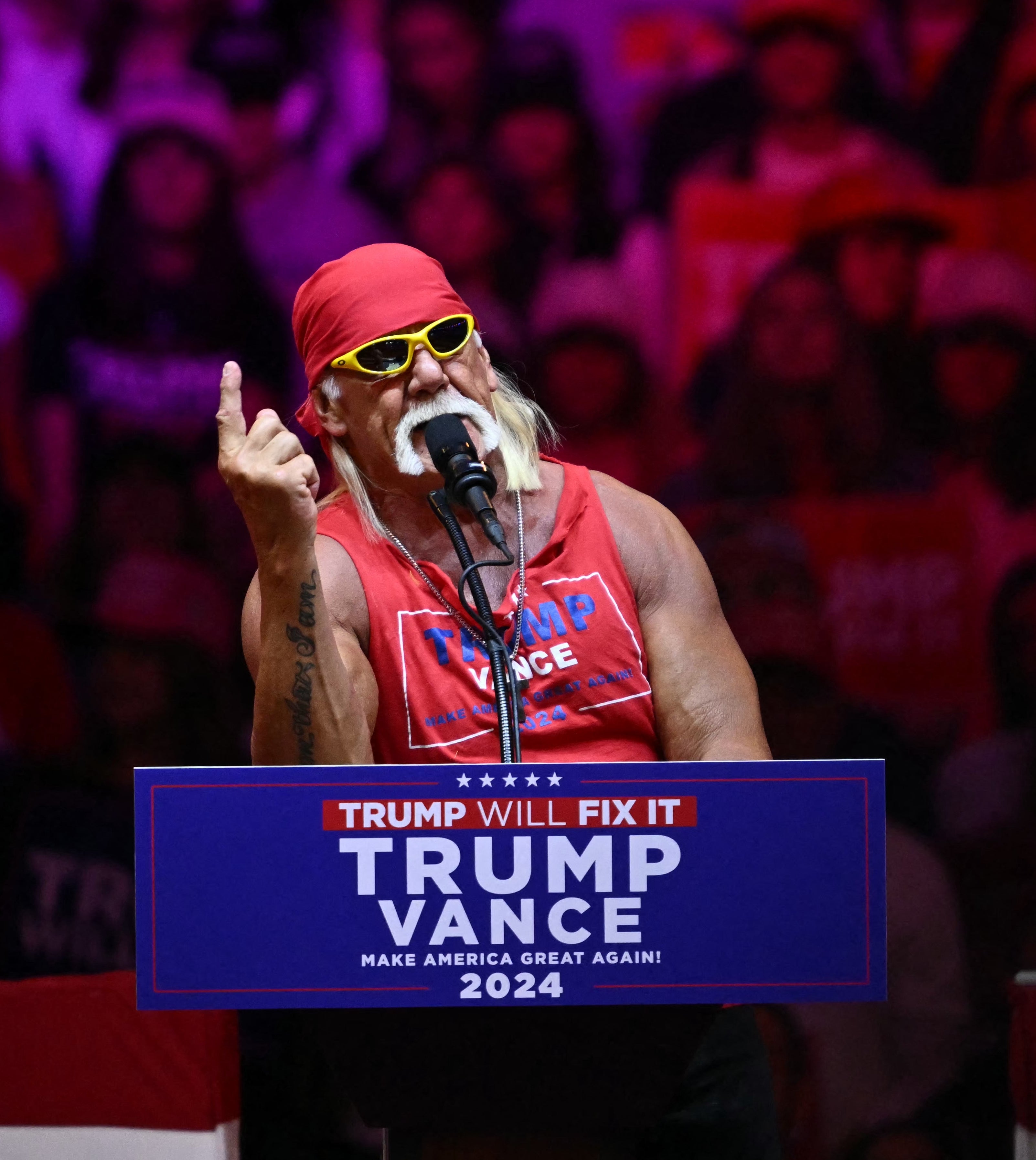 A person at a rally wears sunglasses and a tank top, speaking at a podium with &quot;Trump Vance 2024&quot; signage. Audience visible in the background