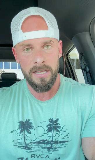 Person with a beard and a cap is sitting in a car, wearing a T-shirt with palm trees and sunset graphic
