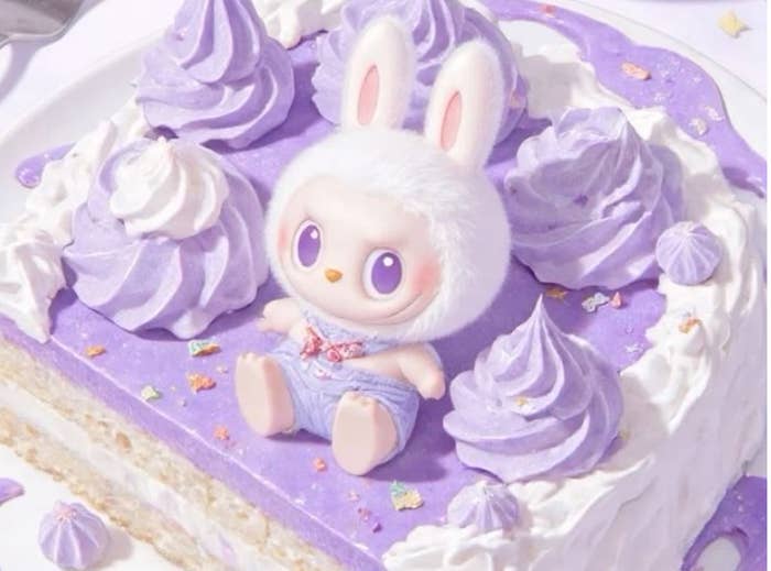 Muñeco de conejo sentado sobre un pastel decorado con crema y rosetones, tiene una expresión adorable y lleva un atuendo de mezclilla
