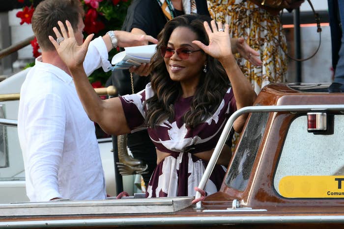 Oprah waves while standing on a boat in stylish attire, featuring a belt and flowing sleeves