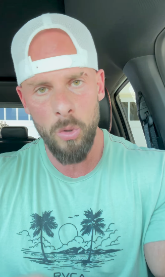 Man with cap and beard talks in a car, wearing a tropical-themed T-shirt