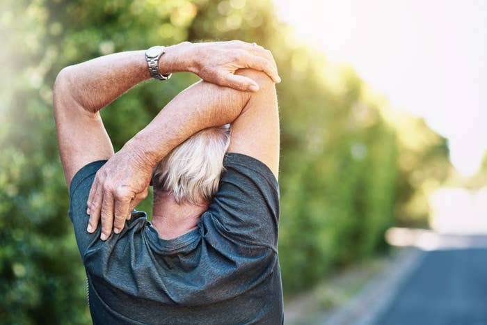 Person stretching outdoors with hands over the head, standing near a path lined with greenery. They are wearing a casual shirt and a watch