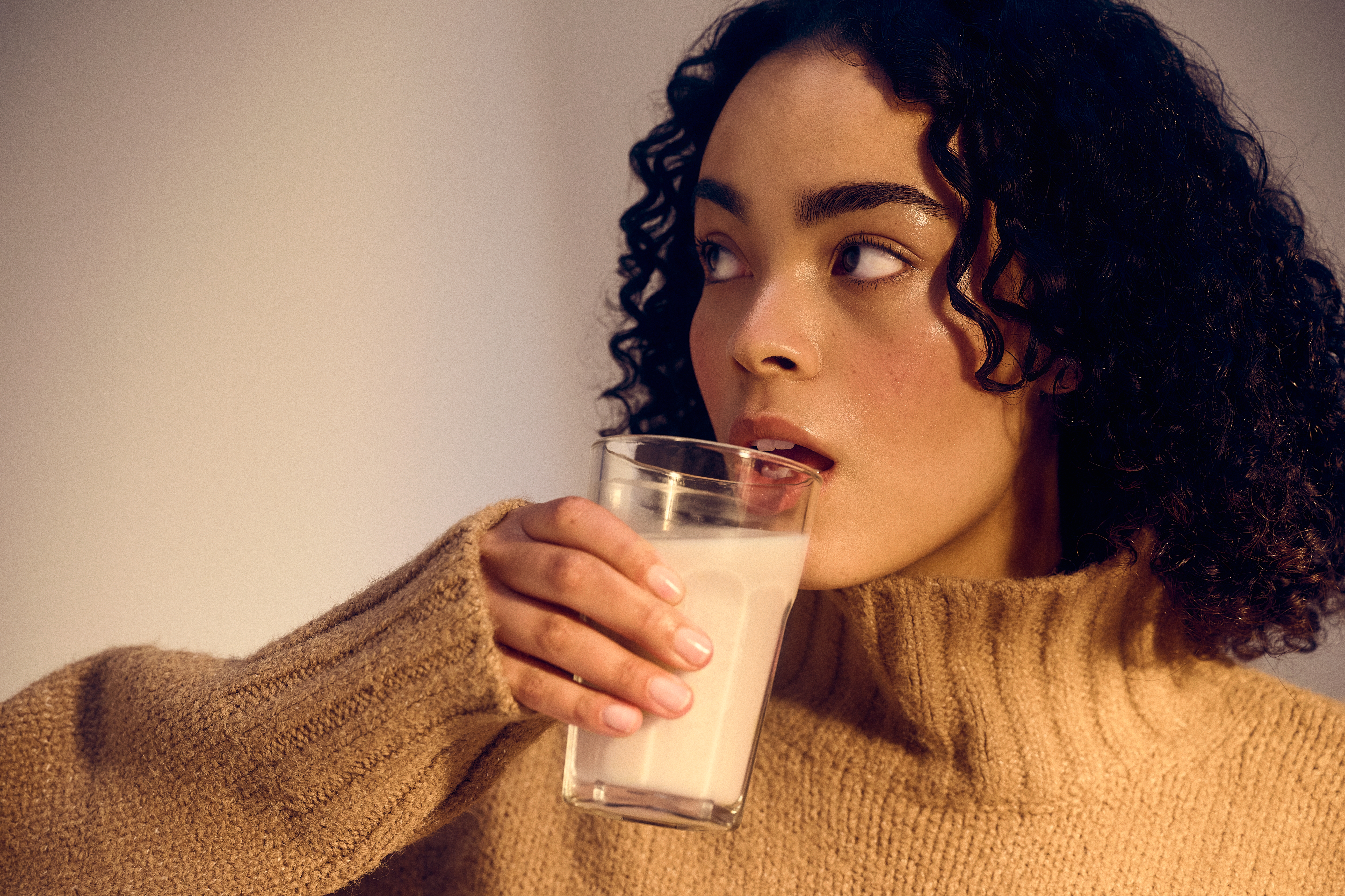 Person with curly hair in a cozy knit sweater drinks from a glass of milk, gazing thoughtfully to the side