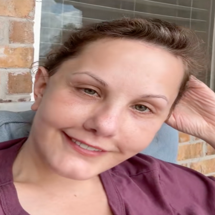 A person smiles at the camera, resting their head on their hand in a relaxed pose against a brick and window backdrop