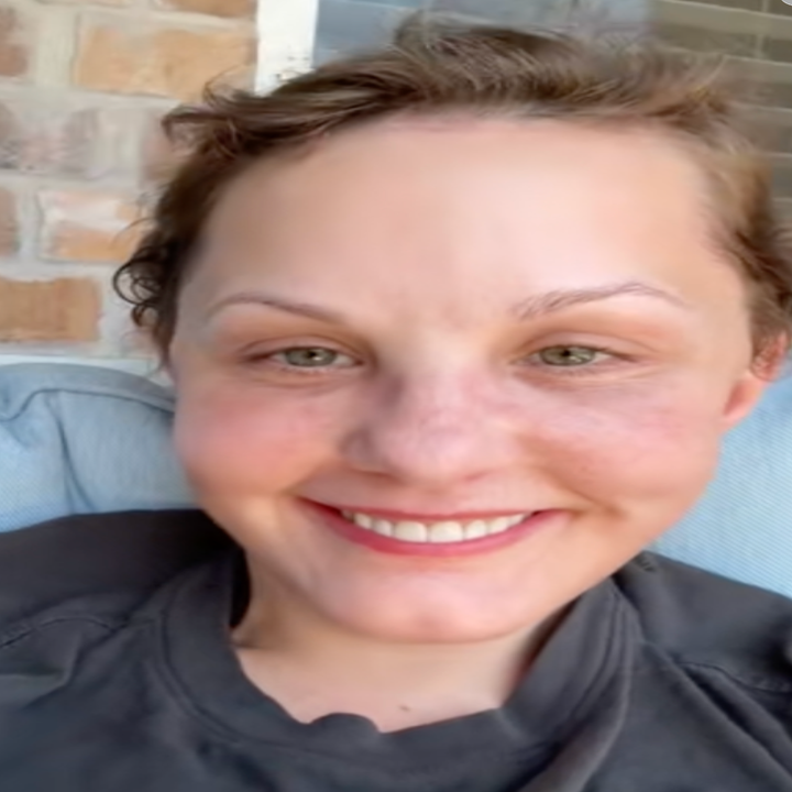 Person smiling while sitting on a couch, wearing a casual t-shirt, appearing relaxed and happy. Background includes a brick wall