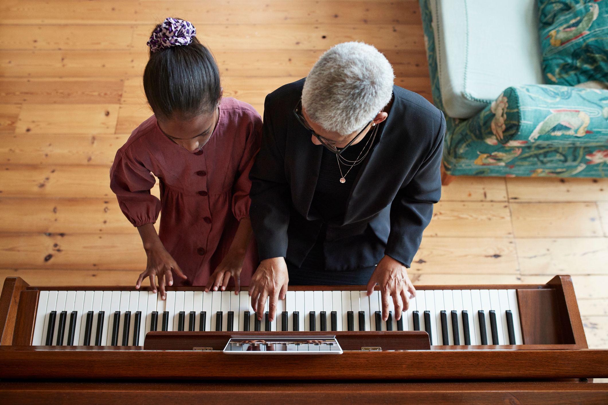 A child in a dress and adult in a blazer play piano together, viewed from above, highlighting a shared musical moment