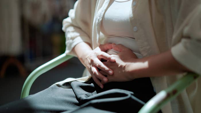 Person sitting on a chair, holding their stomach, suggesting discomfort or contemplation. They wear a light shirt over a white top