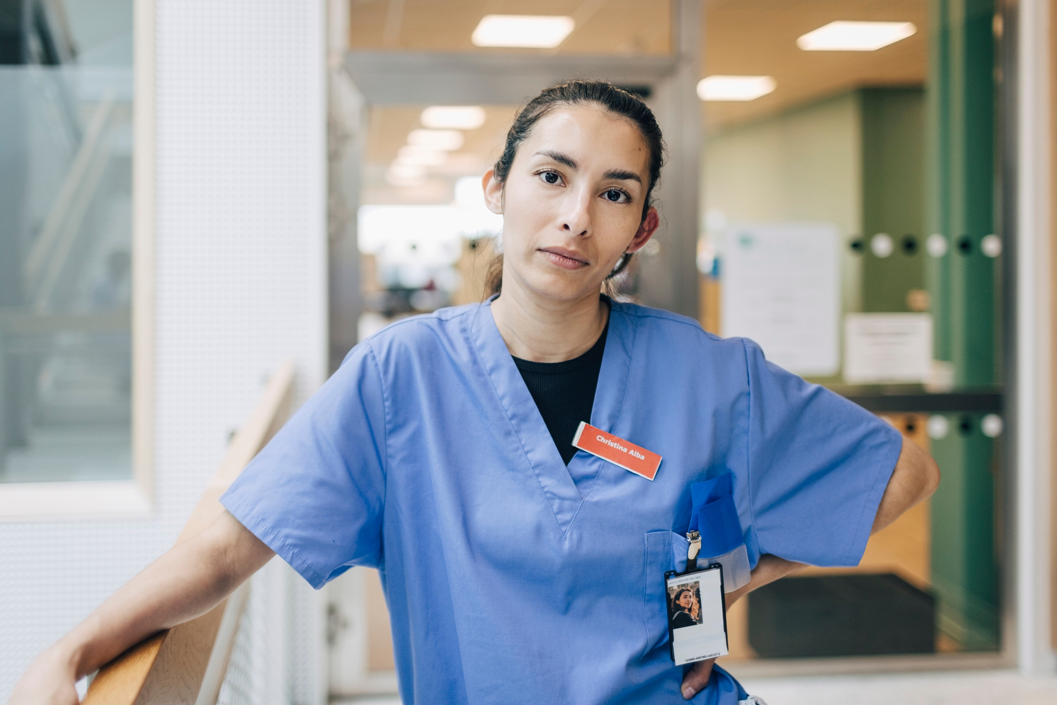 Person in hospital scrubs stands confidently in a medical facility hallway, wearing a staff badge
