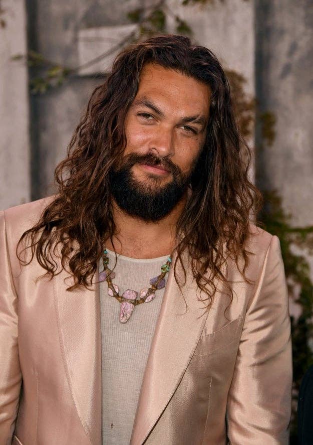 Jason Momoa with long hair and a beard in a stylish suit and necklace