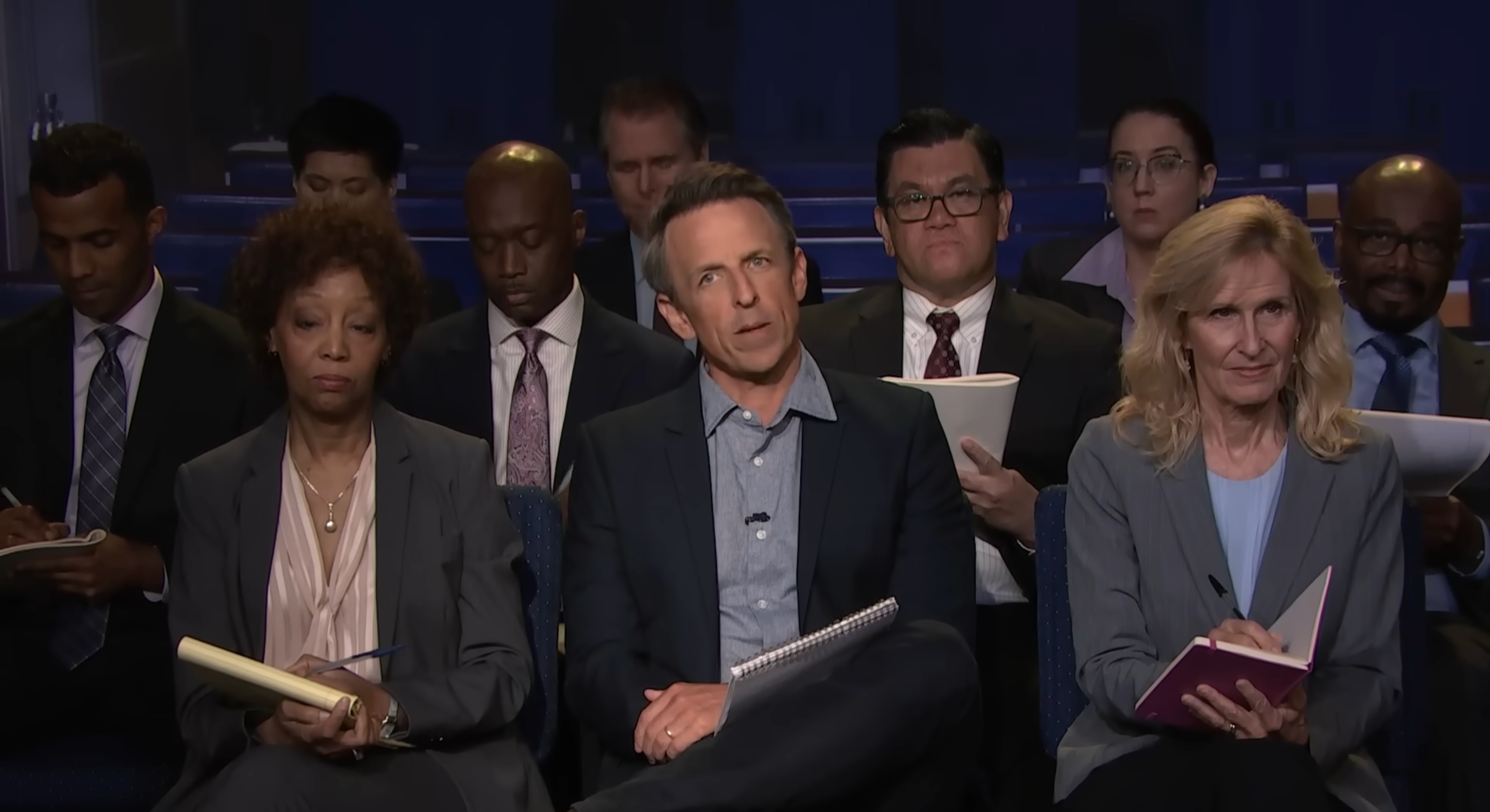 A group of people in business attire, seated and taking notes at a conference or meeting