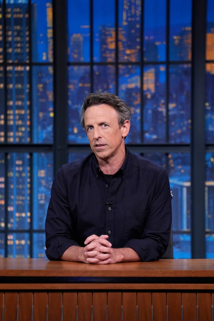 A person sits at a desk with a cityscape backdrop, wearing a casual shirt, appearing to host a talk show