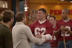 Group of men in casual jeans and matching jerseys in a cafe setting, engaged in conversation, with others watching in the background
