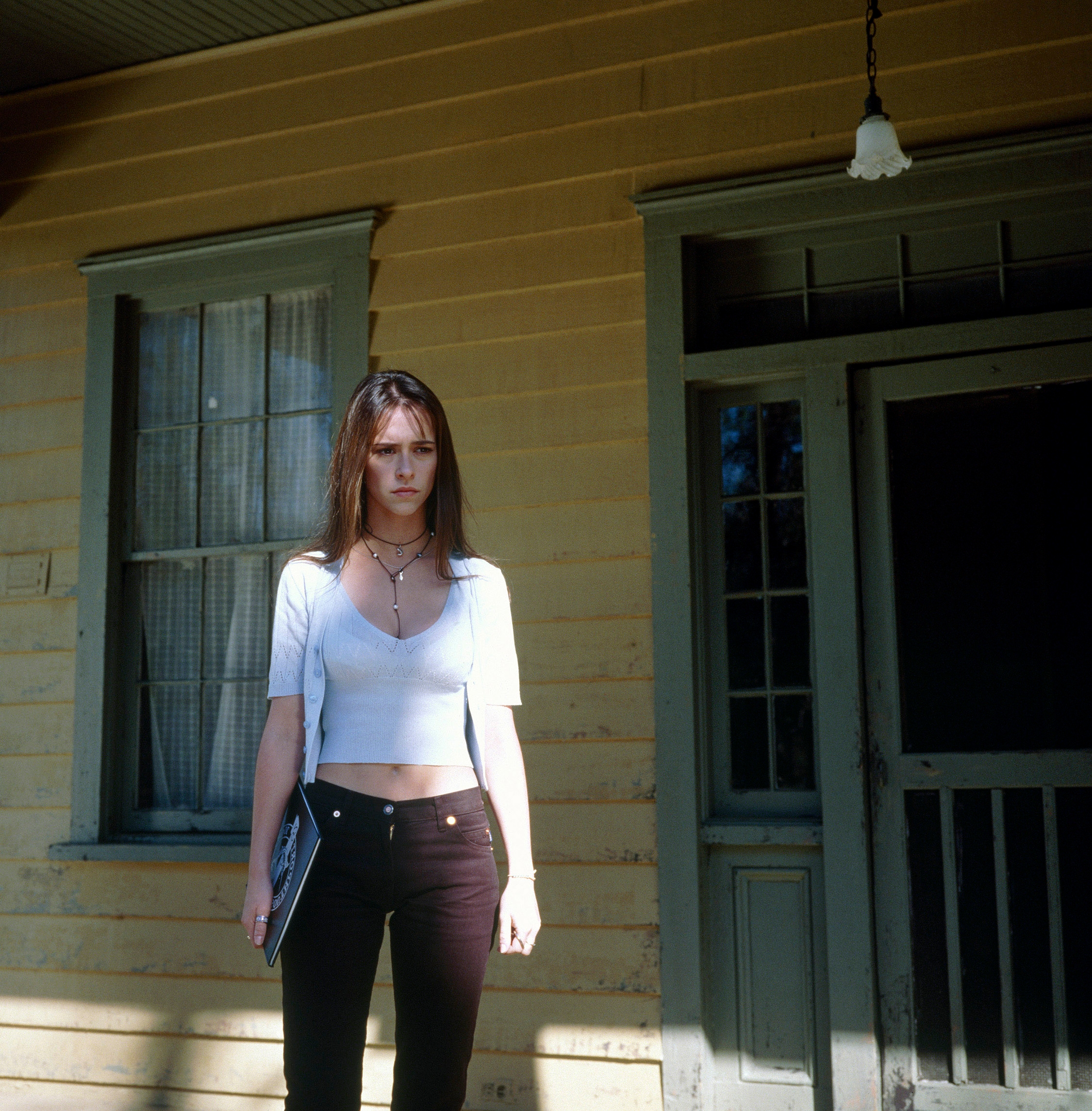 Person stands in front of a wooden house, wearing a white top and dark pants, holding a notebook