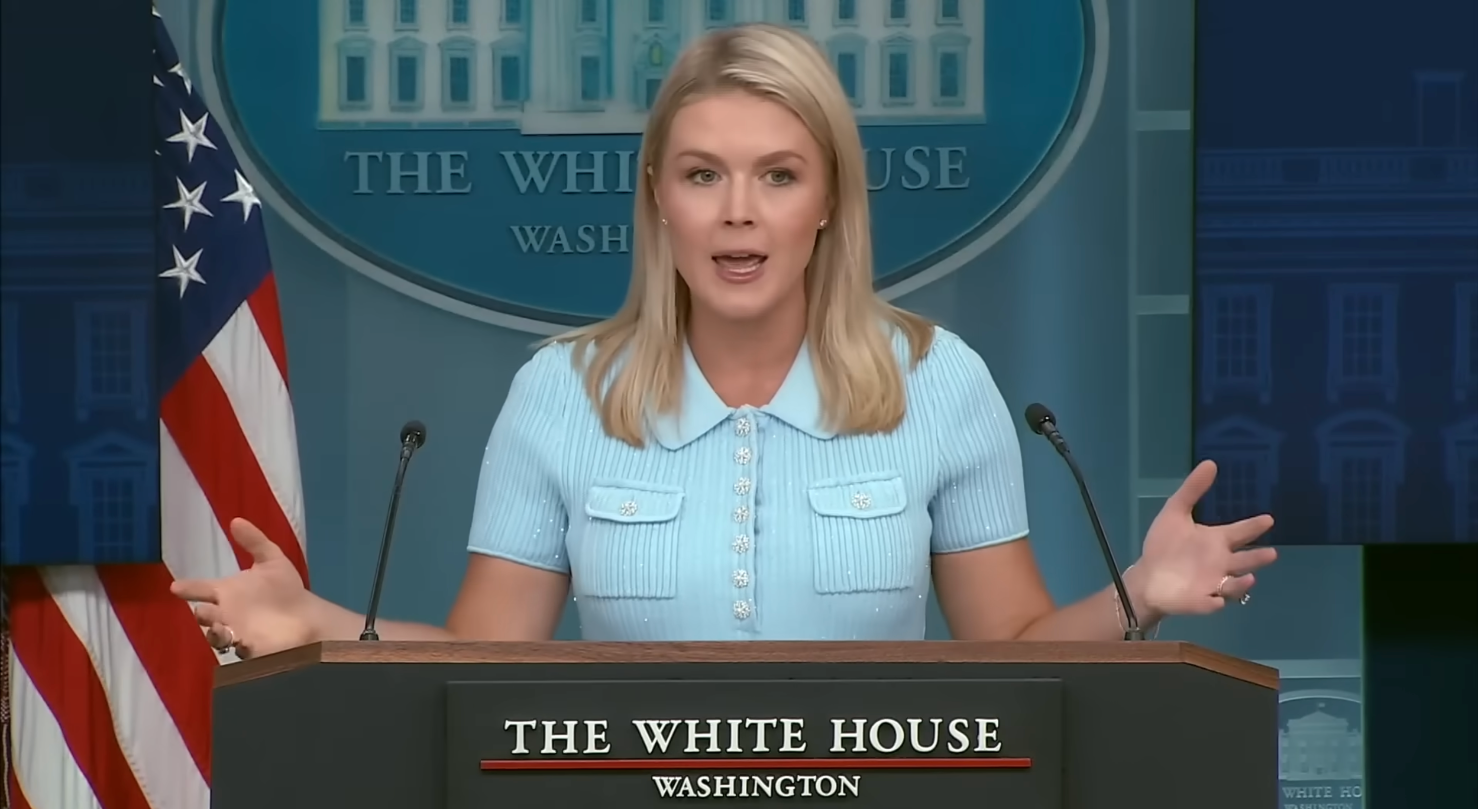 Person speaking at White House press briefing podium, with arms outstretched, in a formal setting