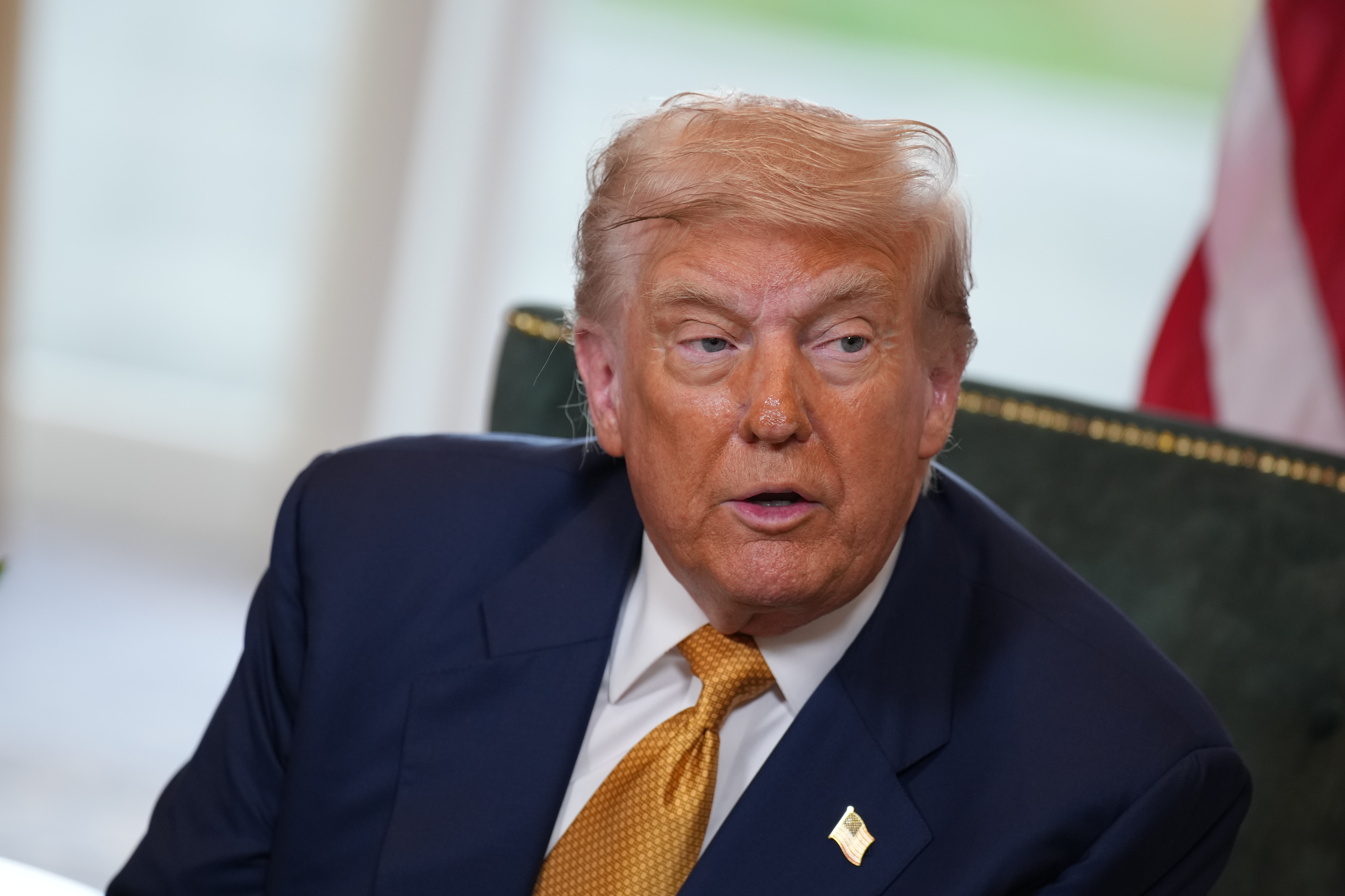 A person in a suit and tie sits, speaking in what appears to be a formal setting, with part of a flag in the background