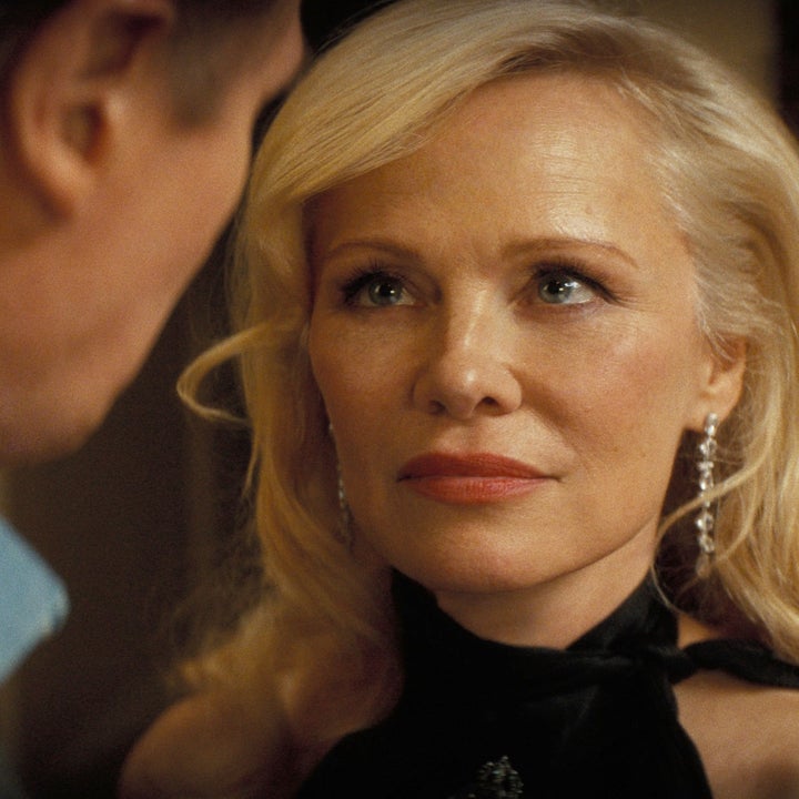 Two people in a close-up scene; one is a woman with wavy hair, wearing a halter top and dangling earrings, looking intently at the other person