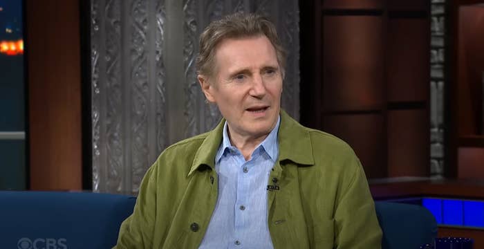 An older man in a casual jacket and shirt appears on a talk show set. He is engaged in conversation, sitting on a blue couch