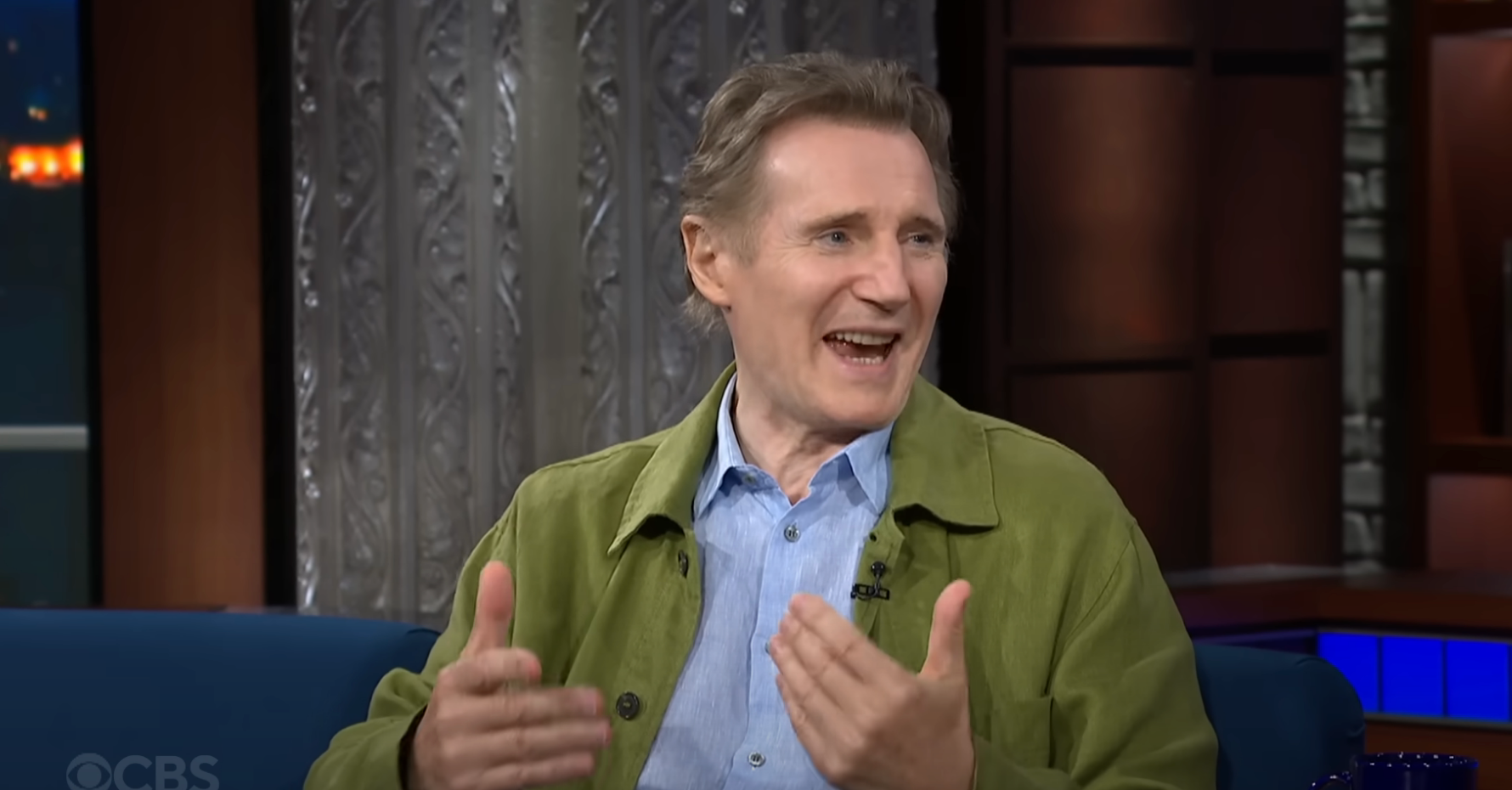 Man in casual jacket gestures during a talk show appearance. He's animated, with a backdrop of a studio setting