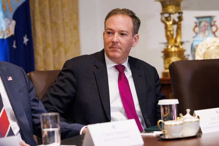 A man in a business suit with a purple tie sits at a conference table with documents and a teapot in front of him