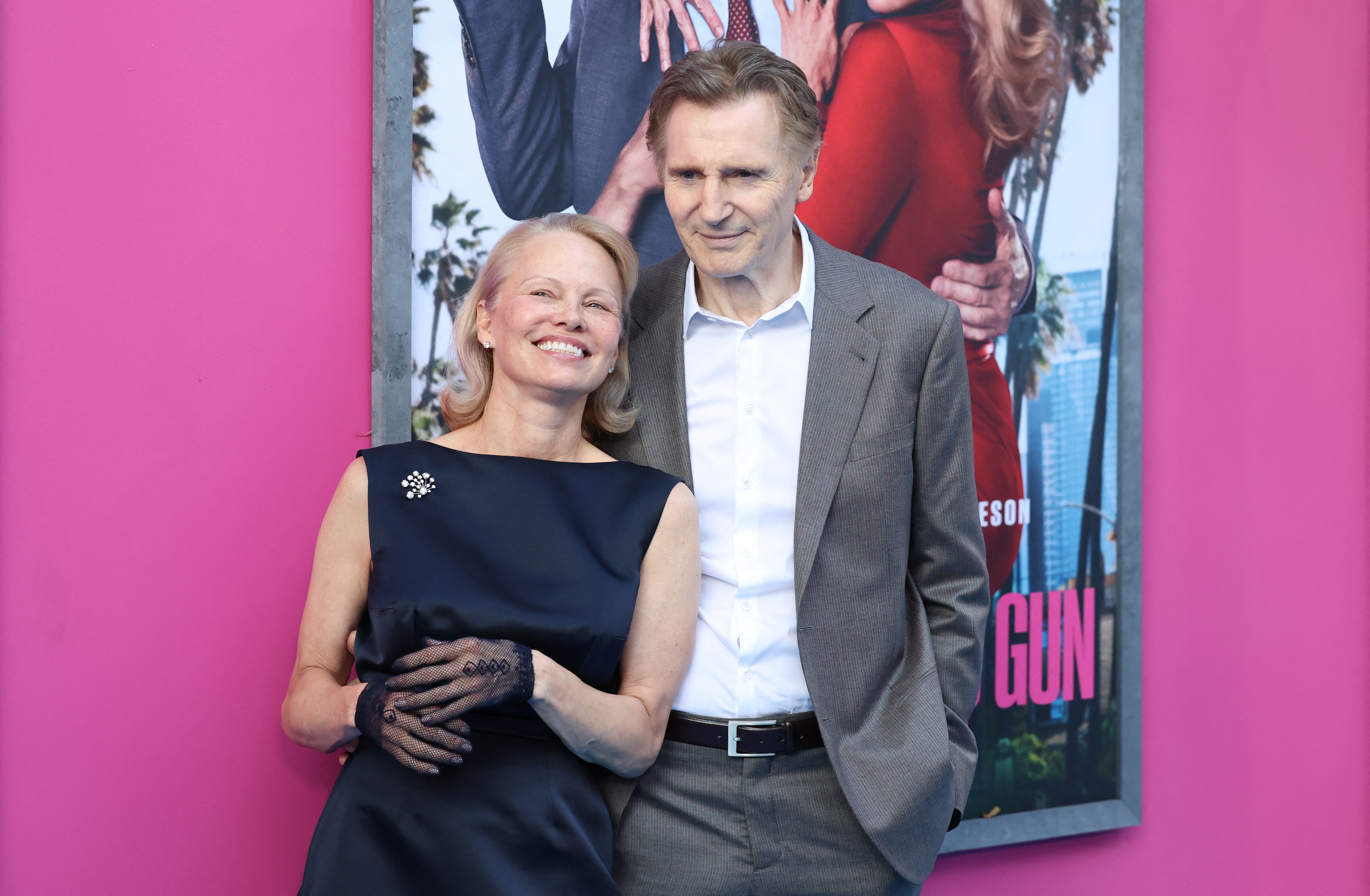 A man and woman pose in front of a movie poster at an event. The woman is in a dark sleeveless dress with lace gloves; the man wears a gray suit