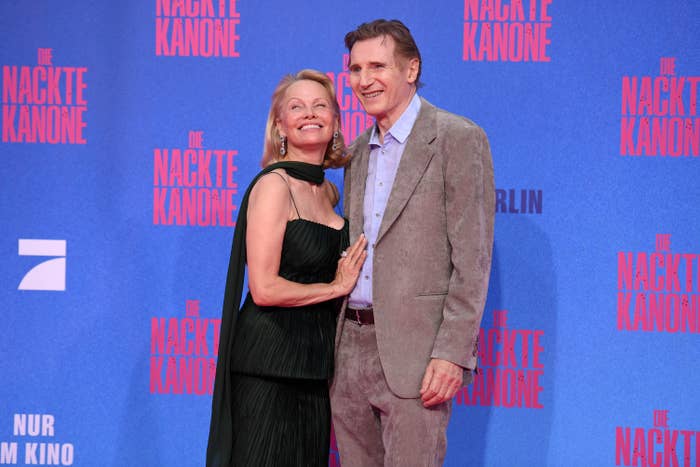 Two people pose on a red carpet. One wears a strapless, pleated dress with a shawl; the other is in a suit without a tie, both smiling