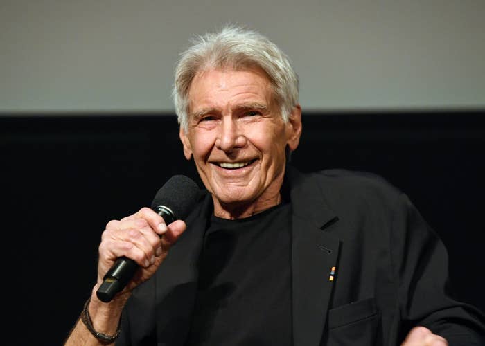 A man in a black jacket and shirt, smiling while holding a microphone during an event