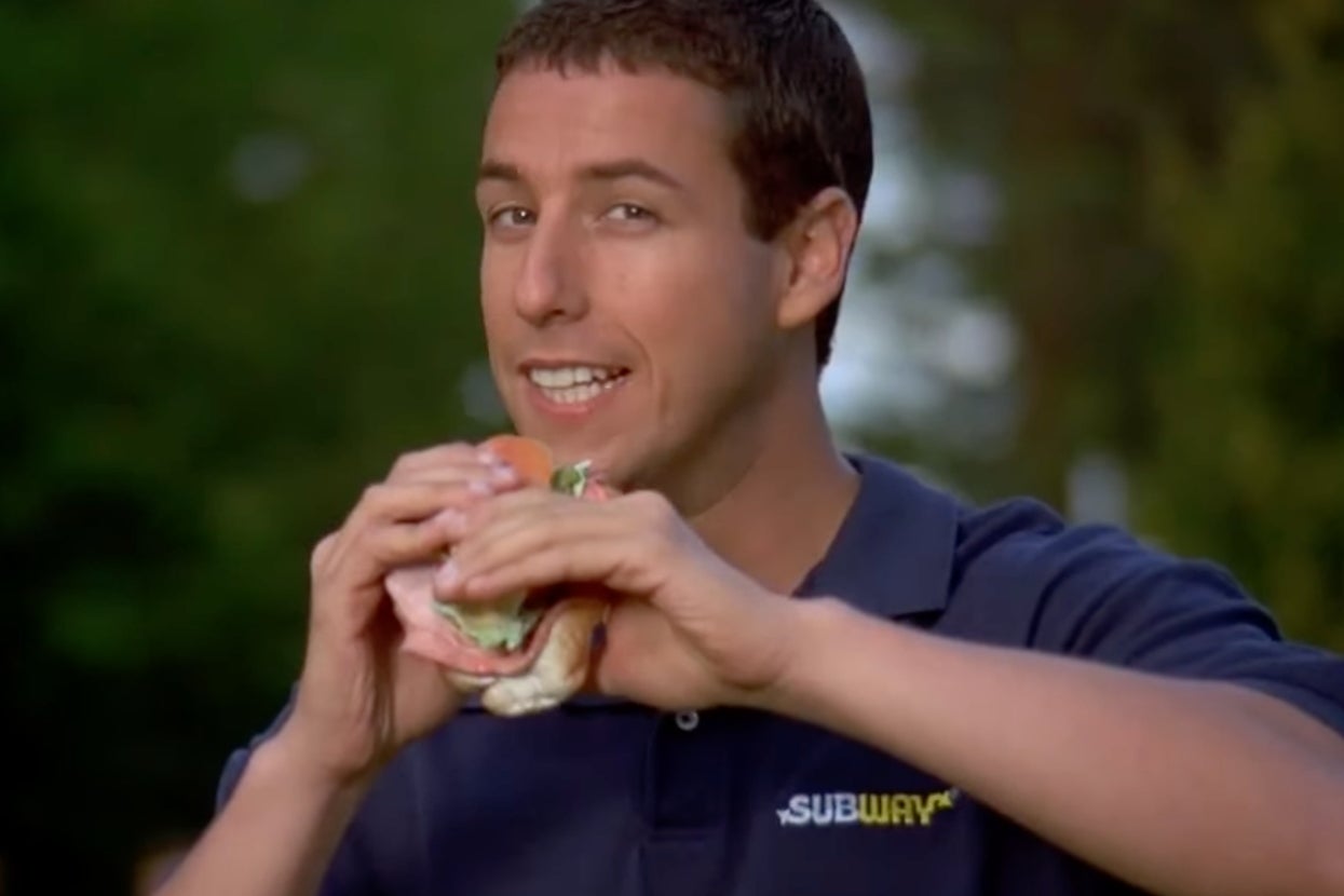 Adam Sandler in "Happy Gilmore" in a Subway shirt holds a sandwich, smiling and about to take a bite, outside in a park-like setting