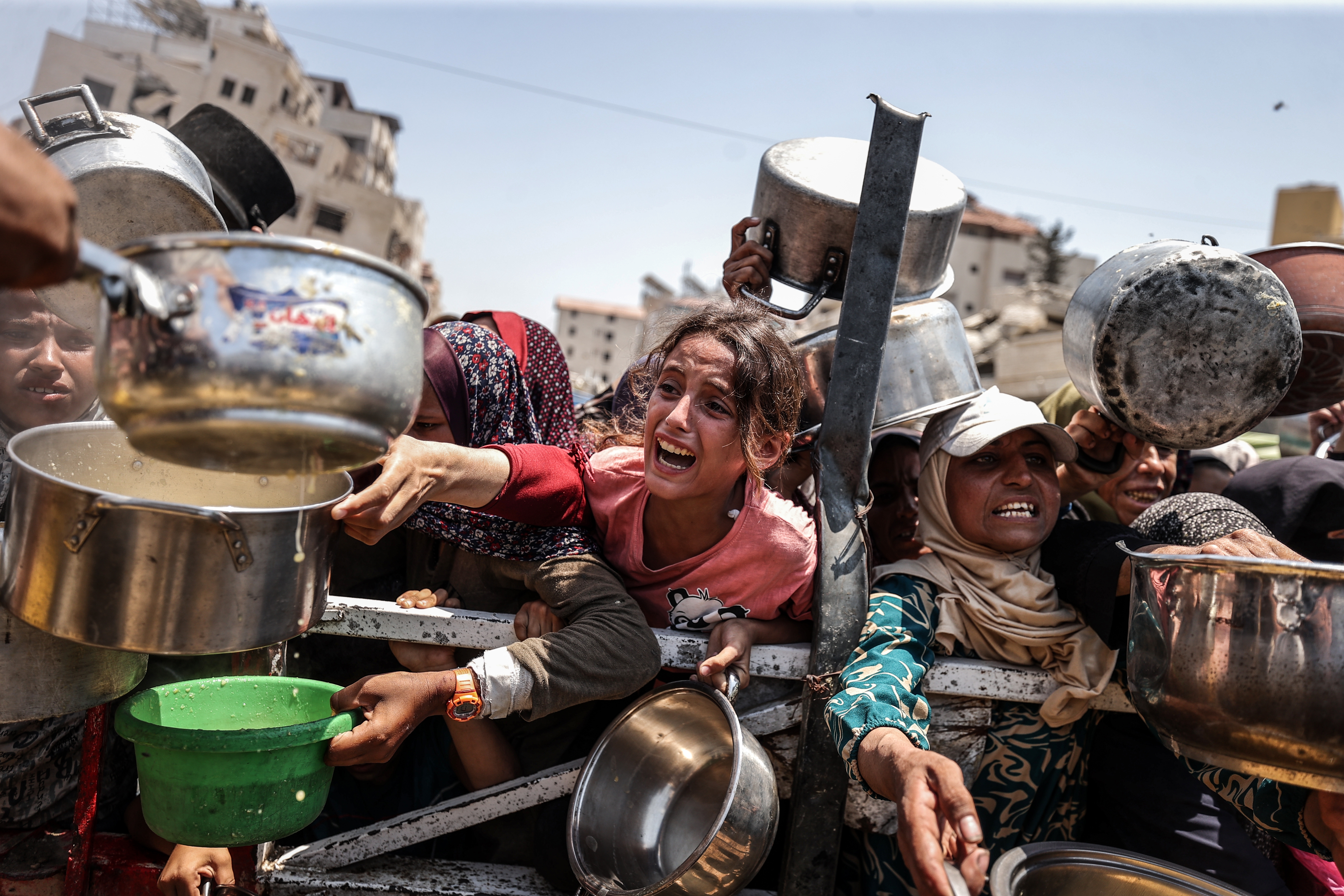 People reaching over a barrier, holding pots and containers. A woman in the center appears distressed, with others around her stretching for aid