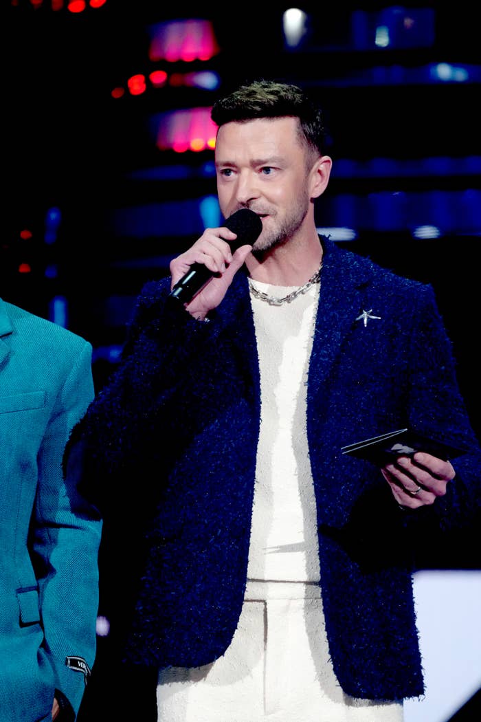 Justin Timberlake in a textured suit holds a microphone, presenting on stage at a public event