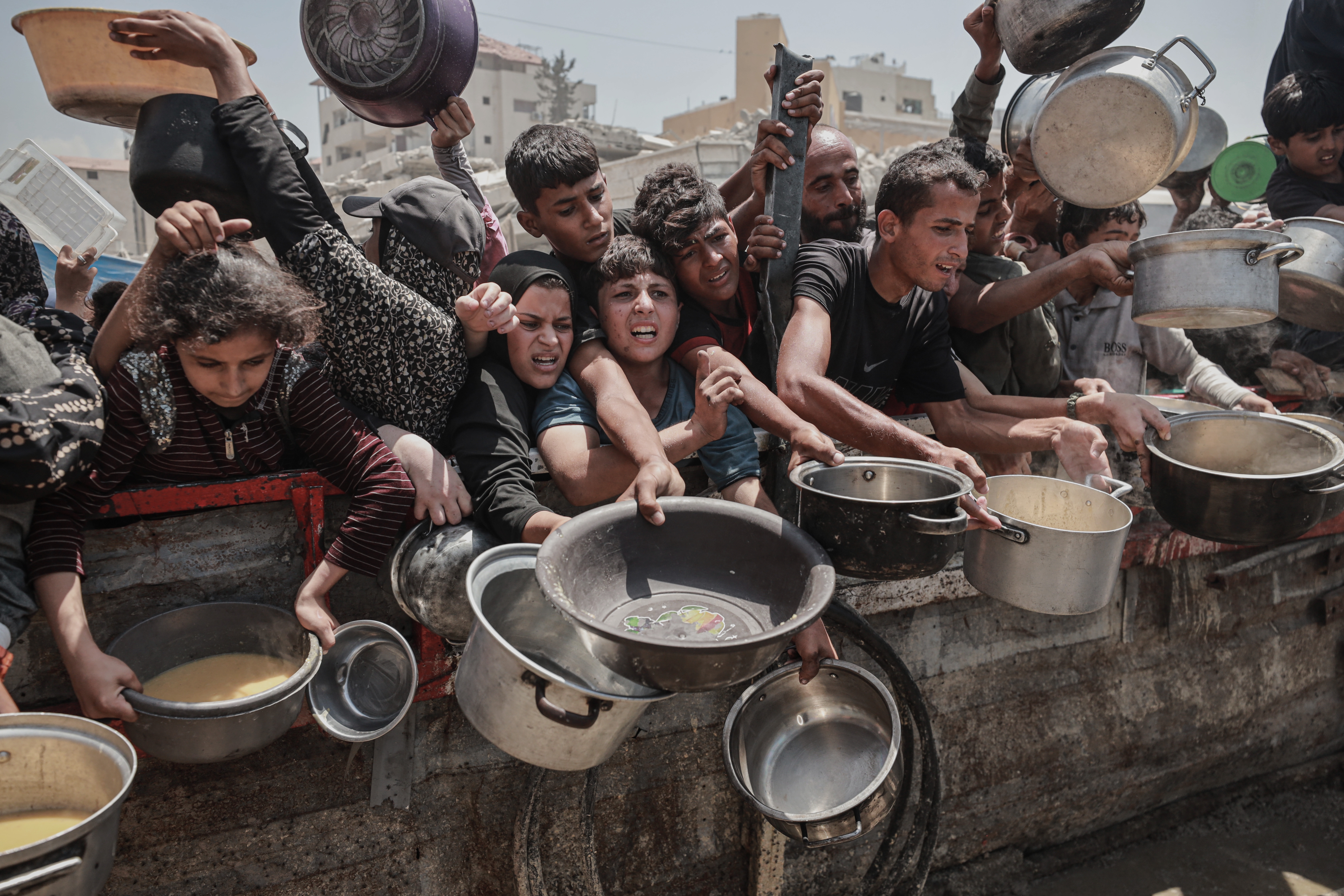 People in crowded conditions holding pots and bowls, appearing to be in a queue for food distribution