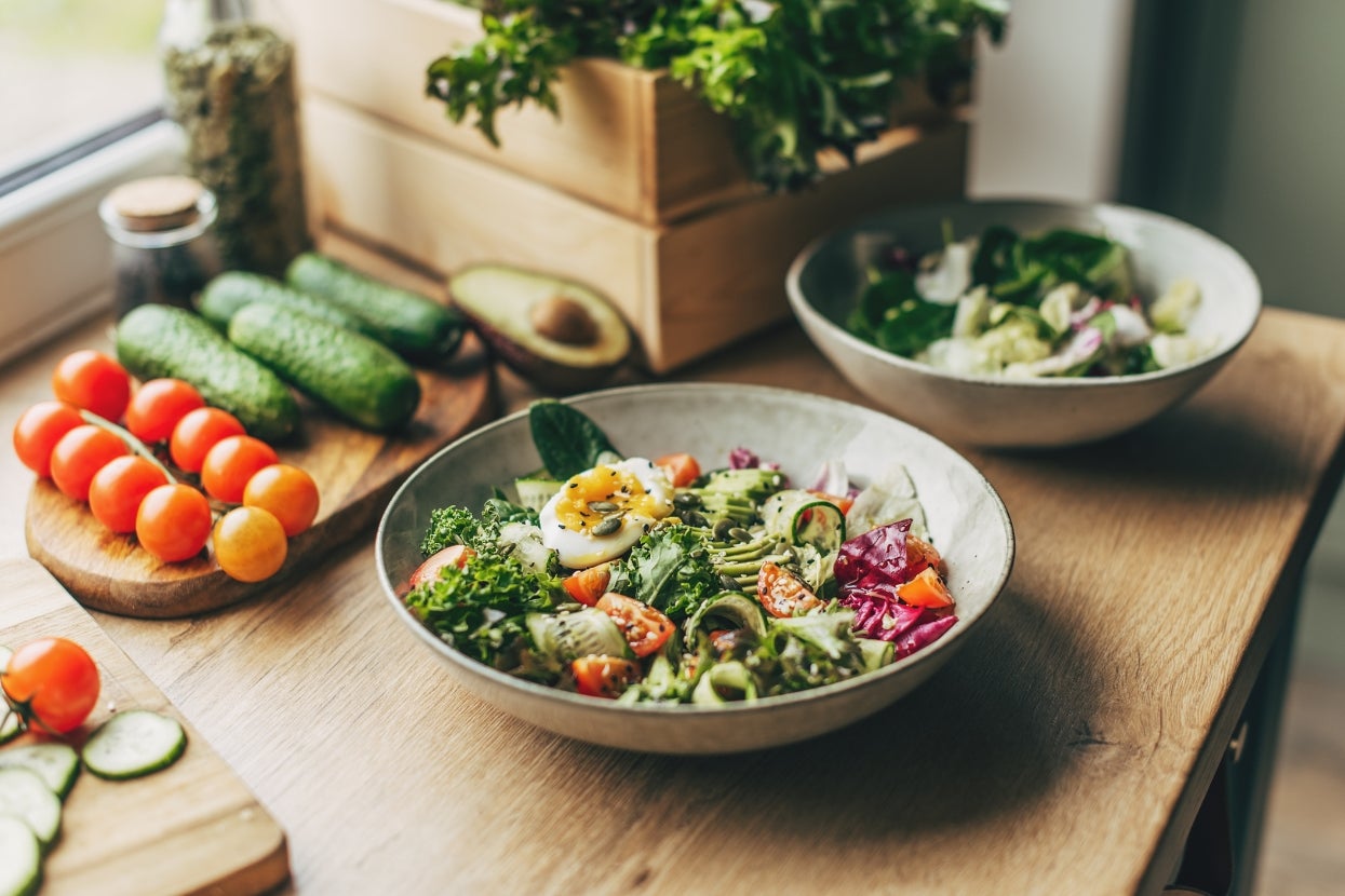 Two bowls of fresh salad with greens, tomatoes, cucumbers, and avocados on a wooden table near a window with natural light