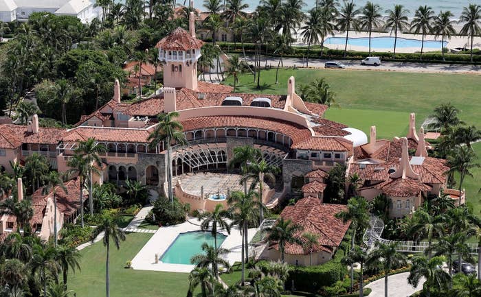 Aerial view of a large estate with red-tiled roofs, palm trees, and a swimming pool, surrounded by greenery and overlooking a beach
