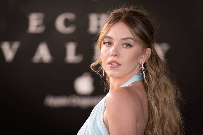 Person in elegant halter dress on the red carpet, looking over shoulder with earrings visible; letters blurred in background