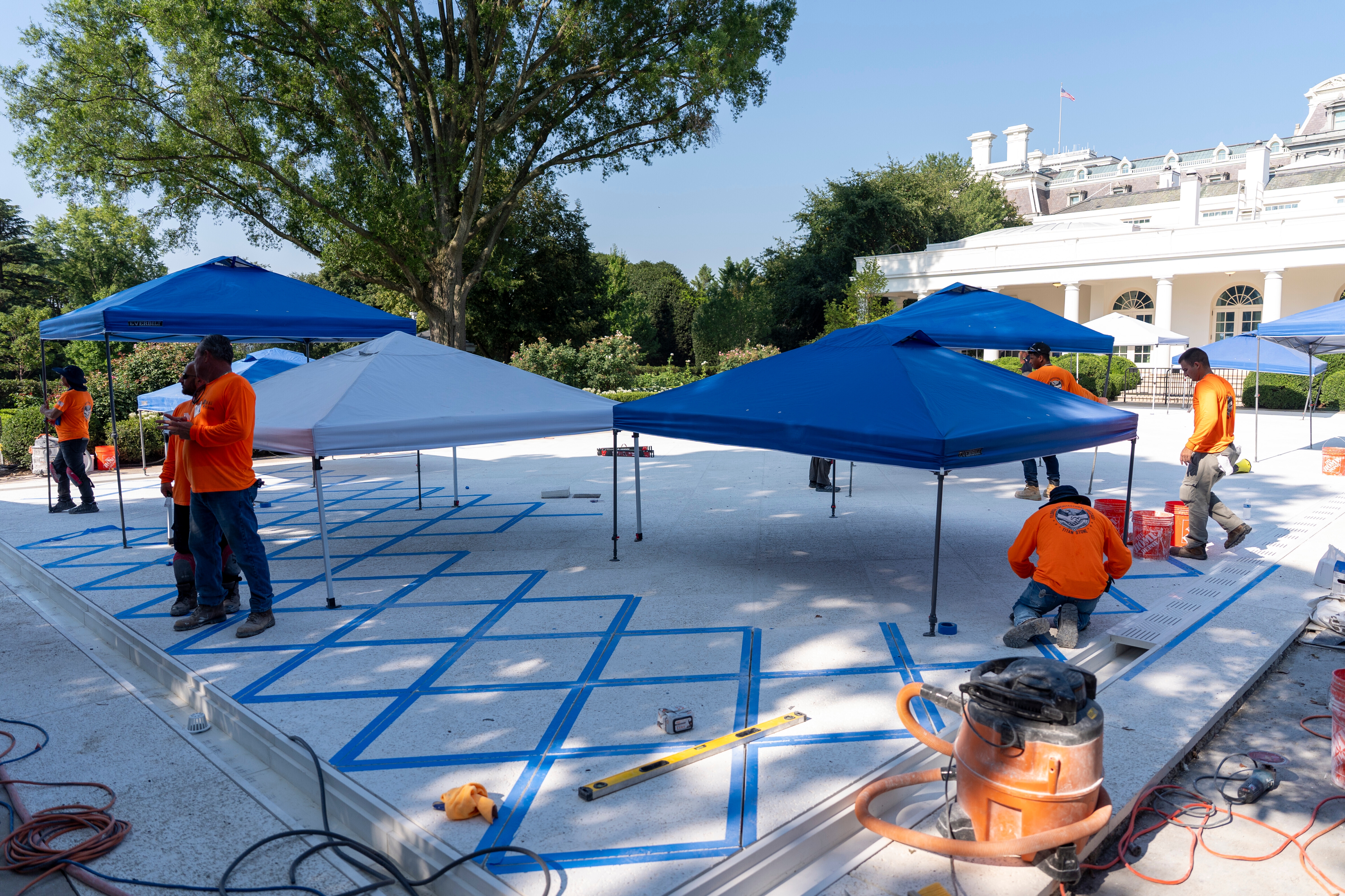 Workers setting up blue and white canopies on a large outdoor platform, with trees and a white building in the background