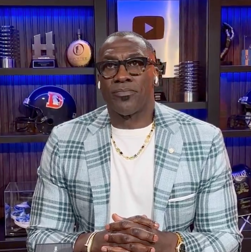 Man in checked blazer and white shirt sits with hands folded, in a room with sports memorabilia on shelves behind him