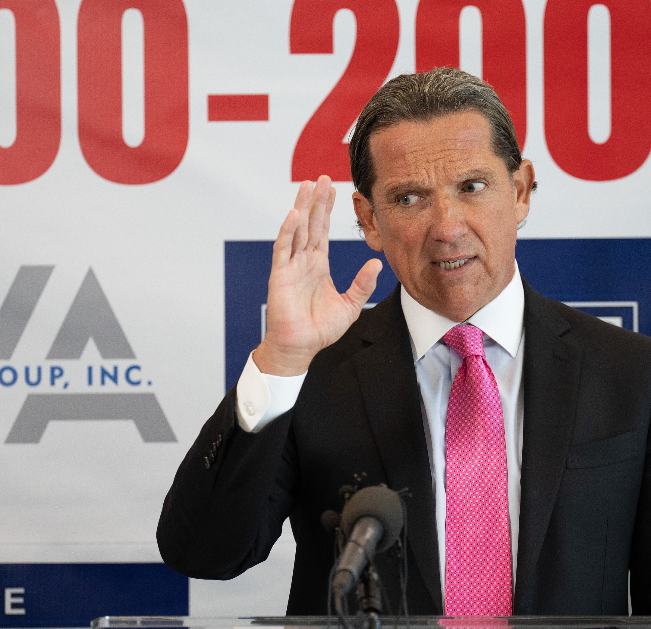 A person in a suit gestures while speaking at a press conference. A phone number and law firm name are partially visible on the backdrop