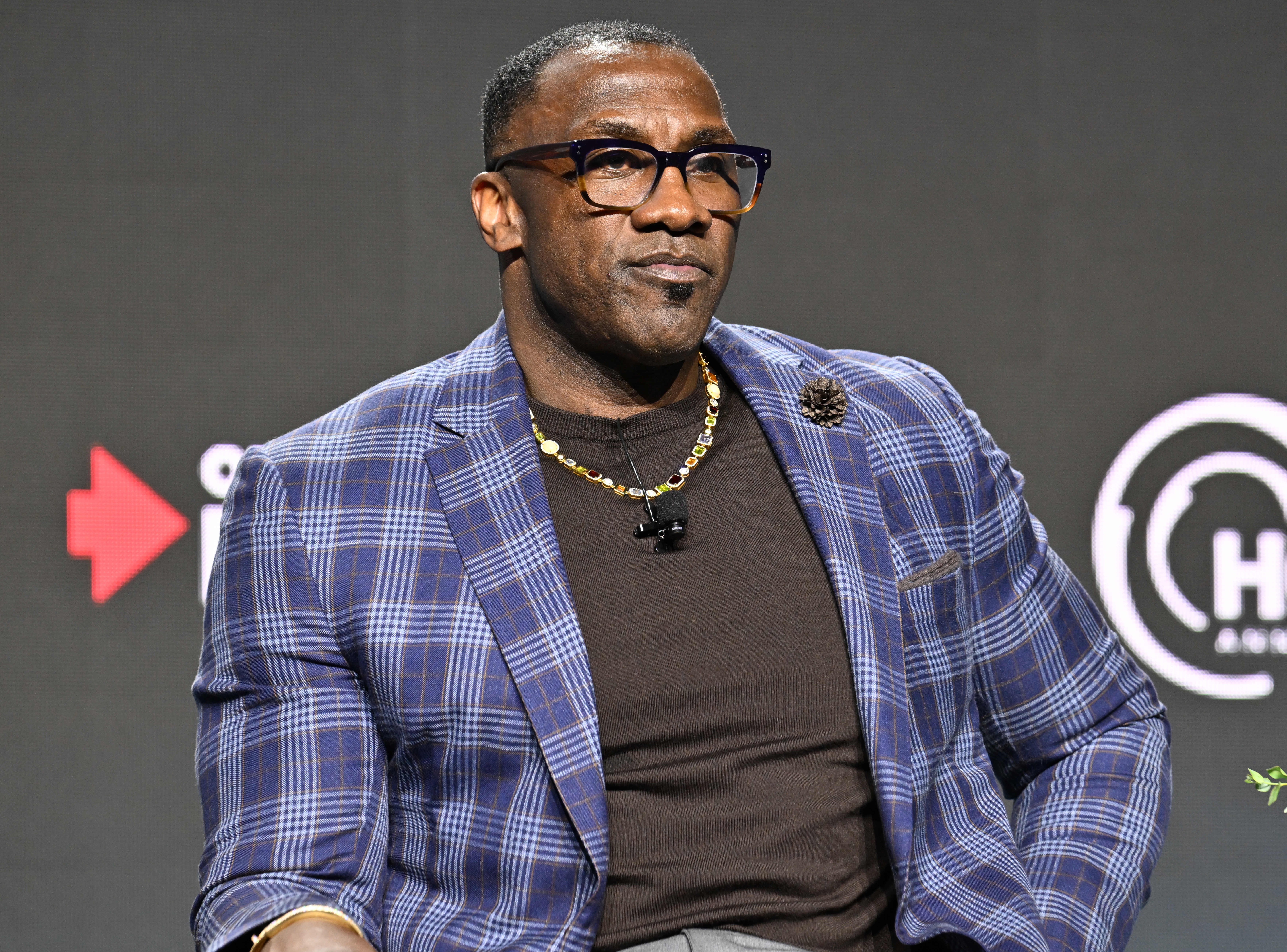 Person in a plaid blazer sits at an event, wearing glasses and a chain necklace, looking thoughtful