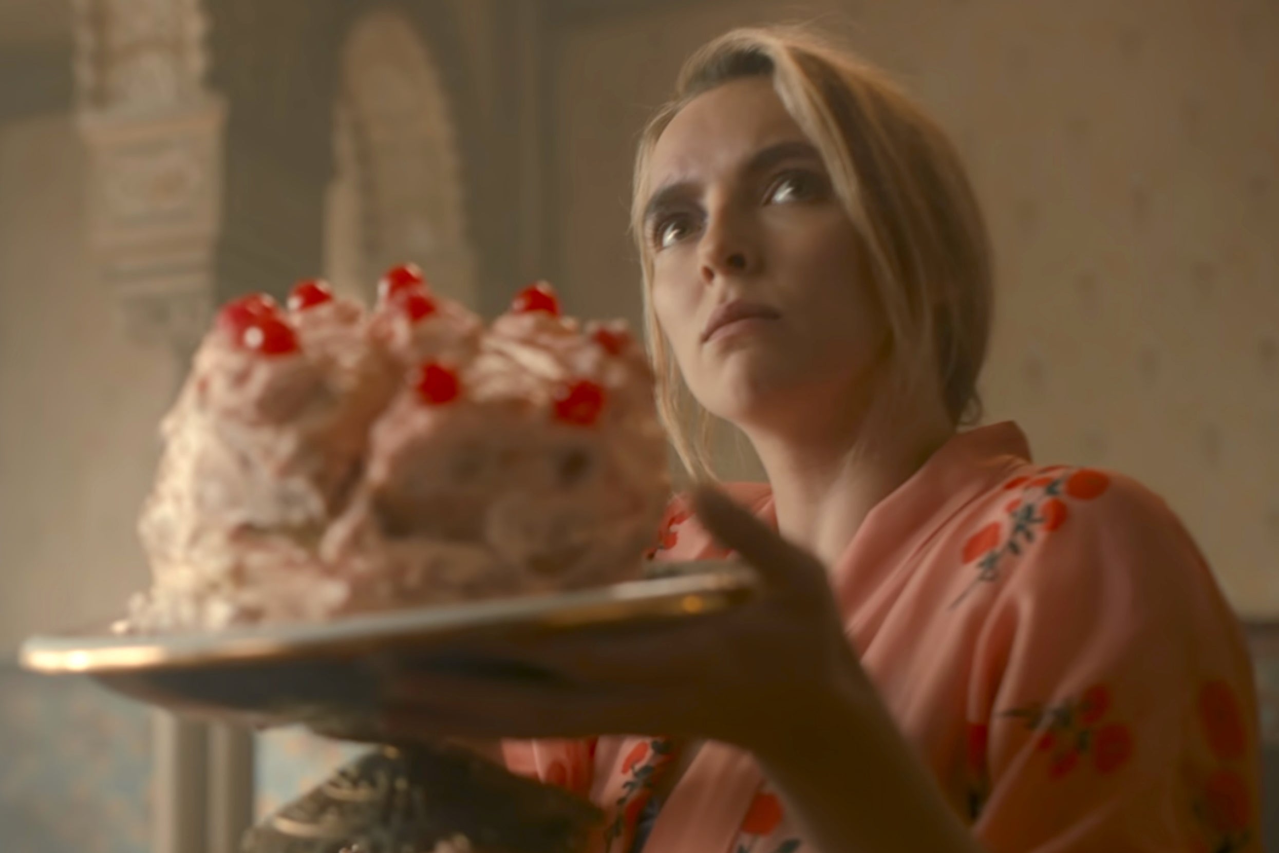 Jodie Comer in a robe holds a cake decorated with cherries, looking upwards with a thoughtful expression as Villanelle on Killing Eve