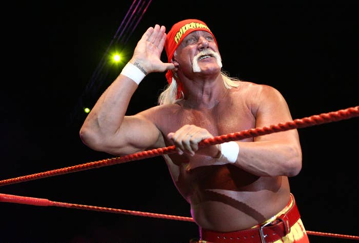 Wrestler in a bandana and mustache stands in a ring, cupping ear toward the crowd, wearing a tank top and matching belt