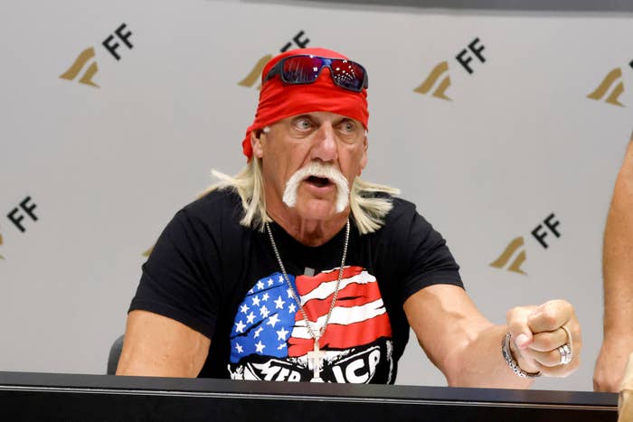 A man in a bandana and sunglasses on his head gestures while sitting at an event table. He's wearing a graphic tee with a patriotic design