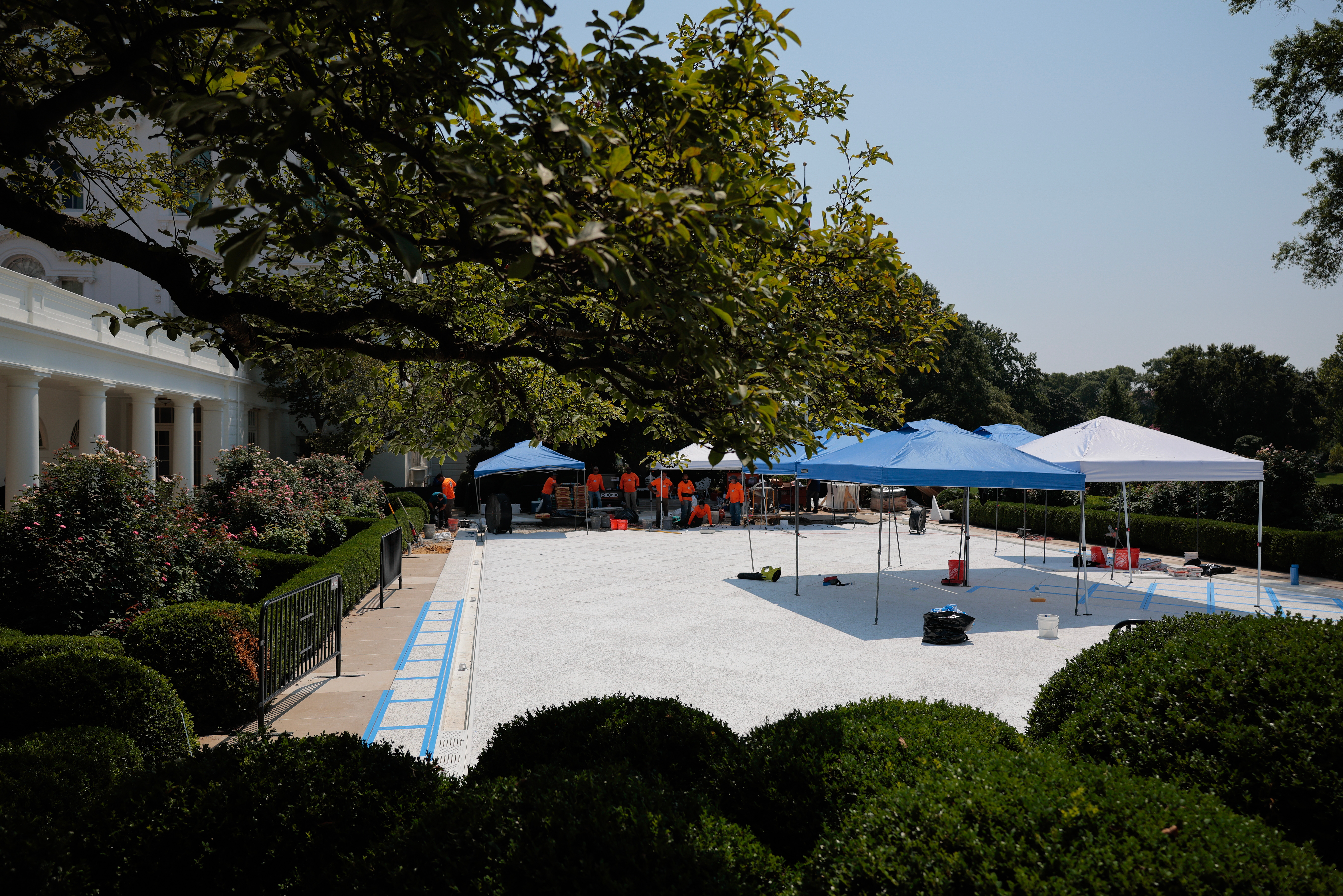 View of a construction project with white tents and workers on a spacious lawn, surrounded by lush trees and greenery