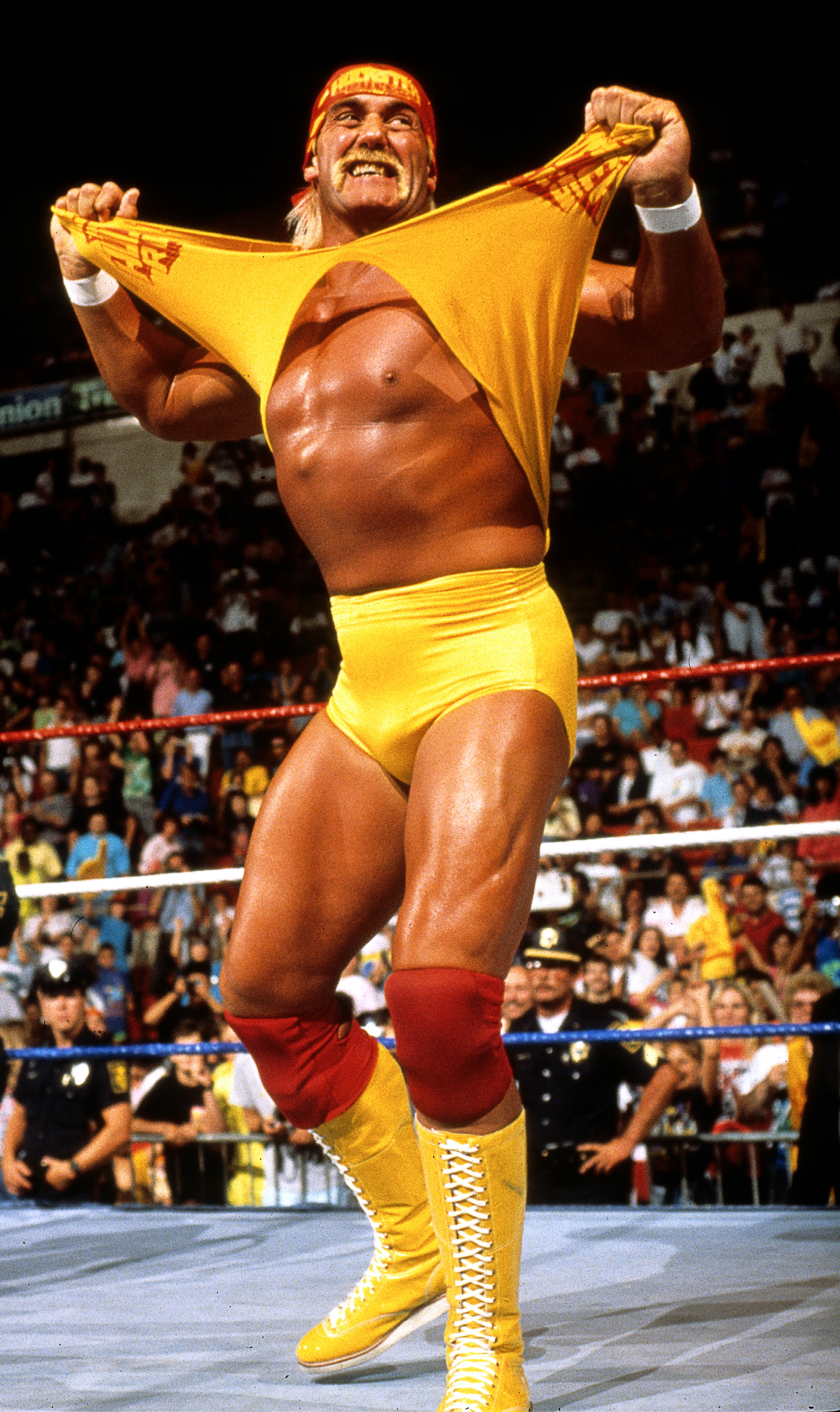 Wrestler tearing tank top in a stadium ring, wearing boots and knee pads, with cheering crowd in background